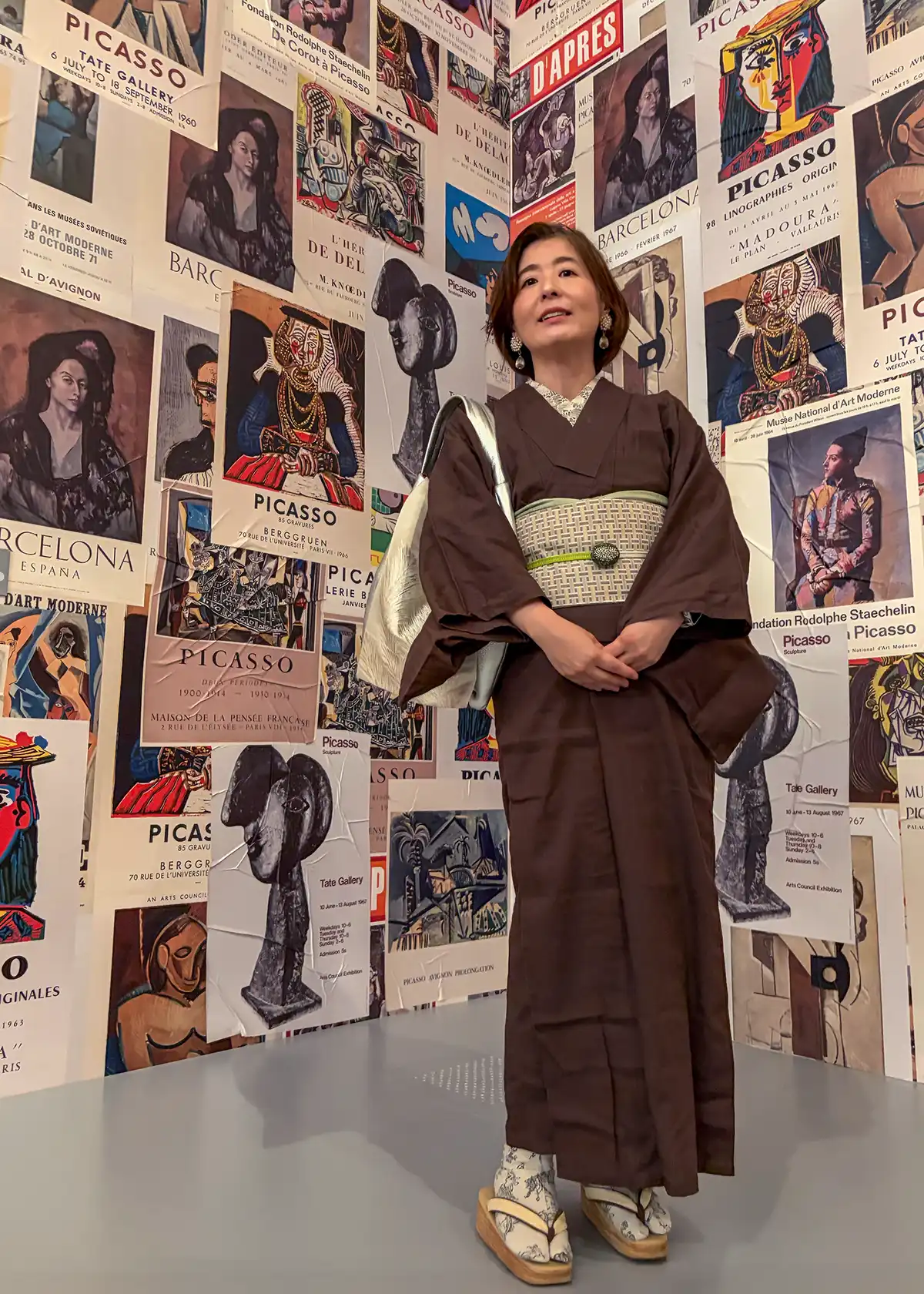歴代のピカソ展のヴィンテージポスターが壁一面にランダムに敷き詰められた空間。その前で、シックなチョコ色のコットン着物に幾何学模様の帯を締めた女性が佇む全身スナップ