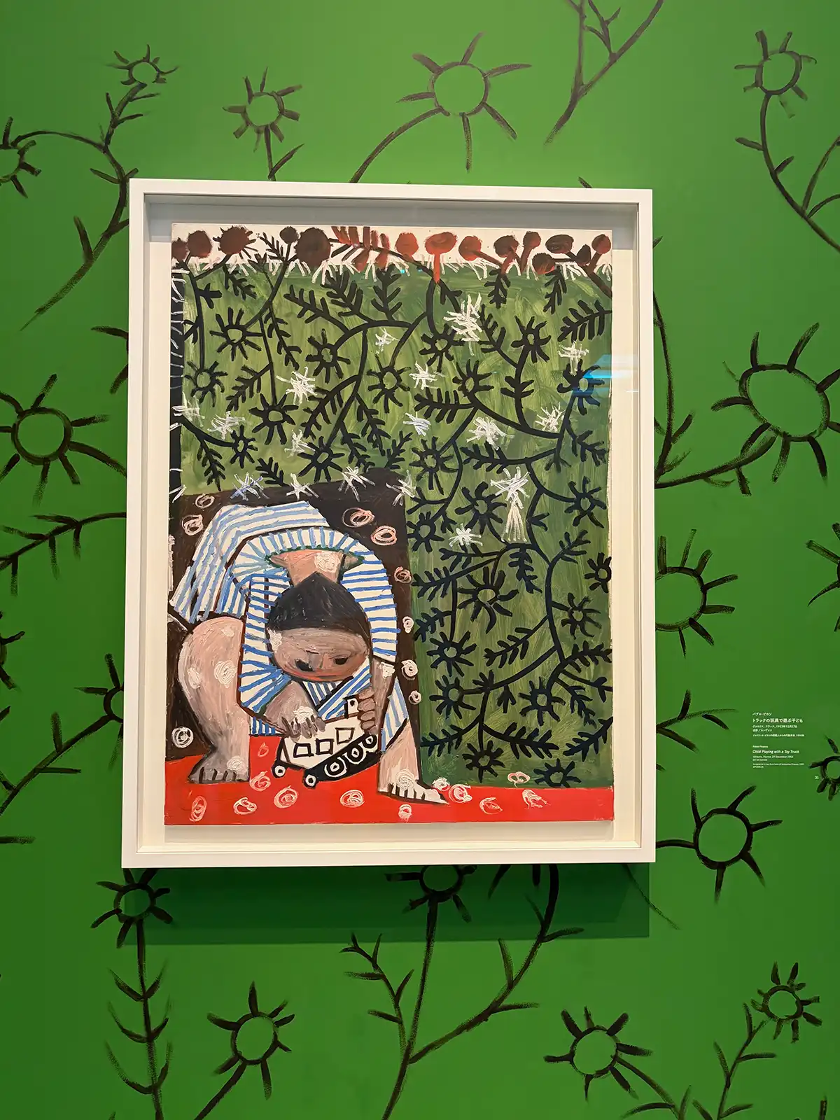 ストライプ柄の服を着た子どもを描いたピカソの絵画《トラックの玩具で遊ぶ子ども》。背景の緑色の壁には、絵の中の植物をオマージュした黒い線画の落書きが大胆に描かれている展示演出