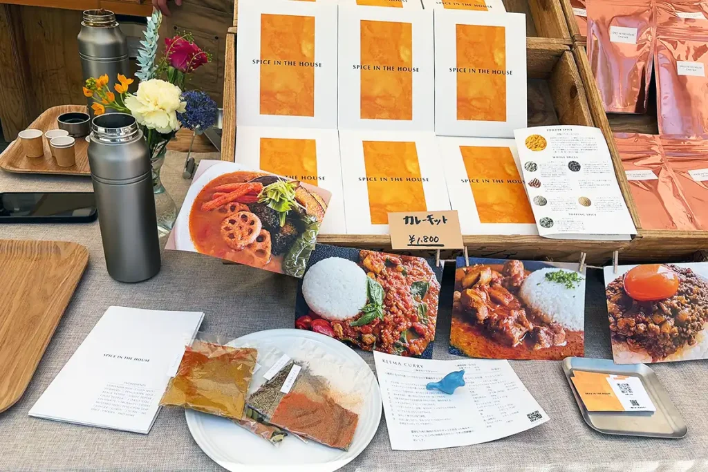 カレー専門店「SPICE IN THE HOUSE」のスパイスキットと、おいしそうな数種類のカレーの写真が展示されたテーブル。
